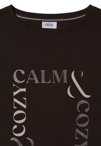 Brązowa koszulka z okrągłym dekoltem, z metalicznym srebrnym napisem "CALM & COZY" w artystycznym układzie. Miękka tkanina o gładkiej fakturze.