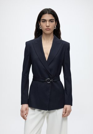 Femme aux longs cheveux foncés porte un blazer sombre à fines rayures avec ceinture et un pantalon blanc, debout devant un fond clair uni.