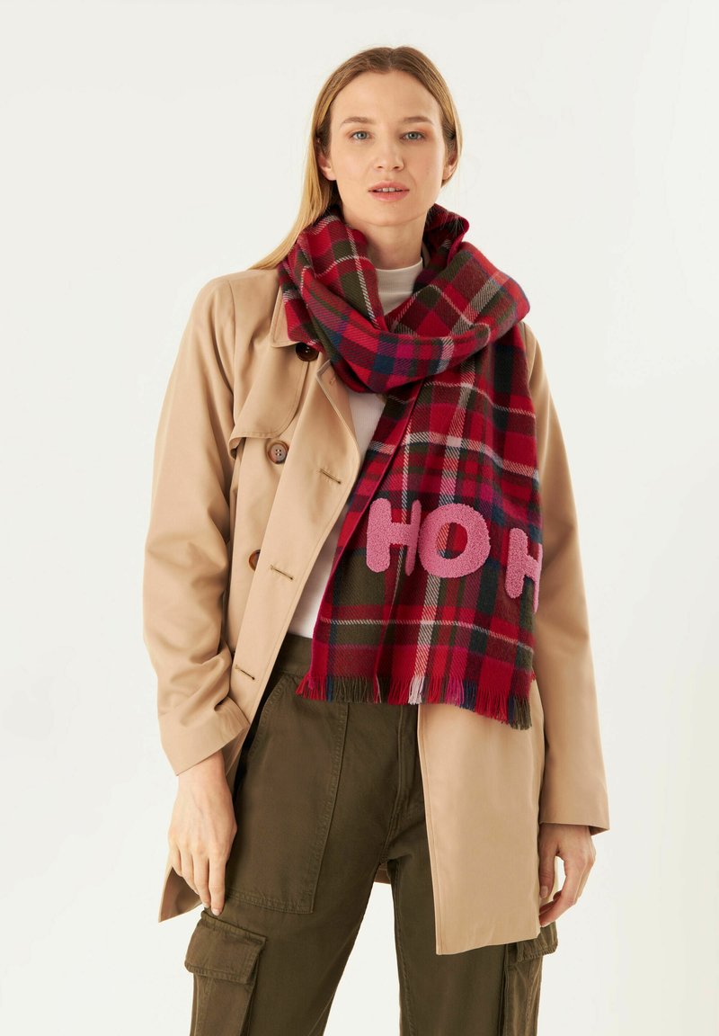 Écharpe à carreaux rouges et verts avec linscription "HO HO" en rose, fabriquée en tissu doux, avec des bords frangés, portée sur un léger pull et un trench beige.