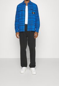 Blue plaid flannel shirt with button-up closure and CK logo, paired with black pants and white sneakers. Focus on check pattern and color contrast.