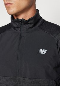 Haut noir de sport à col zippé mi-hauteur avec textures variées, présentant un col haut et un petit logo "N" blanc sur la zone gauche de la poitrine.