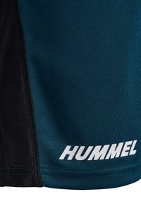 Nærbillede af mørk teal og sort sportstof med hvidt "HUMMEL"-logo trykt nær kanten.