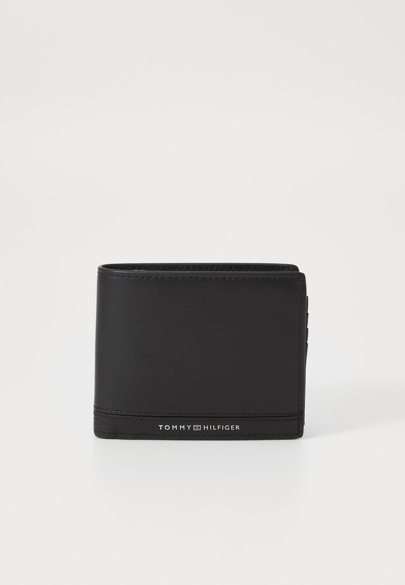 Portefeuille en cuir noir avec une texture lisse, design pliable en deux, et logo en relief "TOMMY HILFIGER" à l'avant. Slim avec des emplacements pour cartes sur le côté.