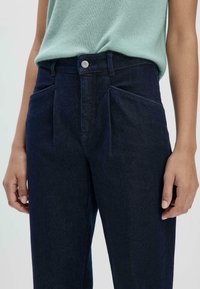 Donkerblauwe hoge taille jeans met geplooide voorkant, schuine zijzakken en metalen knoopsluiting. Glad denim stof met een lichte glans.