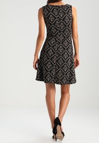 Svart klänning med ett blommönster i damast, utan ärmar, figursydd liv, utsvängd kjol, textil med struktur, och kombinerad med svarta högklackade skor.