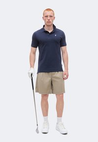 Joven con polo azul marino, pantalones cortos beige, guante y zapatos blancos de golf, sosteniendo un palo de golf, de pie frente a un fondo liso.
