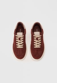 Un paio di sneakers in suede marrone con lacci e suole beige, logo "GUESS" sulla linguetta, mostrato dall'alto su uno sfondo bianco.