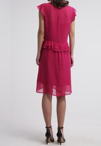 Vestido fucsia con mangas cortas y vaporosas y un volante en la cintura. Tejido transparente, hasta la rodilla, combinado con tacones altos negros.