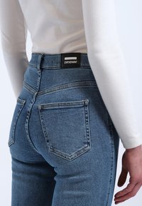 Hochtaillierte blaue Jeans mit glatter Textur, ausgestattet mit einem schwarzen Etikett auf der Rückseite und fünf Taschen mit sichtbaren Nähten.
