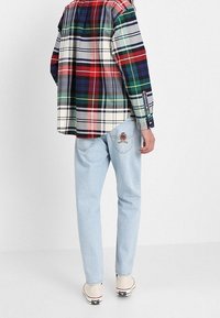 Camicia a quadri con tonalità verdi, rosse e blu, indossata con jeans azzurri chiari con ricami sulla tasca posteriore e scarpe beige.