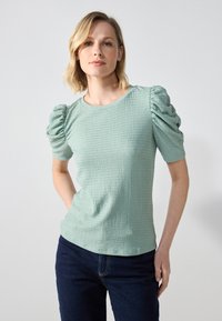 Menta zöld, texturált felső, puffos, ráncolt ujjal, kerek nyakkal és normál fazonnal, sötétkék farmernadrággal párosítva.