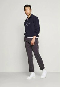Man wearing navy Tommy Hilfiger sweatshirt, dark gray pants, and white high-top sneakers, standing against a plain light background.