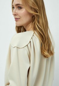 Maglione beige chiaro con dettagli arricciati sulle spalle, morbido al tatto e con un design del colletto arrotondato, mostrato da un lato.
