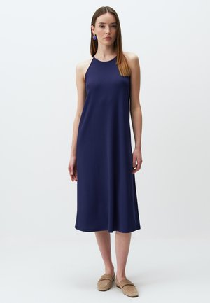 Robe bleu marine à col halter en tissu lisse, avec une coupe décontractée qui tombe sous le genou. Associée à des chaussures légères marron clair à enfiler.