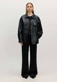 Schwarze Kunstlederjacke mit Knopfverschluss vorne, zwei Brusttaschen und einem lässigen Schnitt, kombiniert mit schwarzen weit geschnittenen Hosen und spitzen Schuhen.