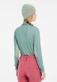 Maglietta a maniche lunghe color teal con colletto alto, pannelli laterali verde chiaro, abbinata a pantaloni rosa con tasca posteriore e vita regolabile.