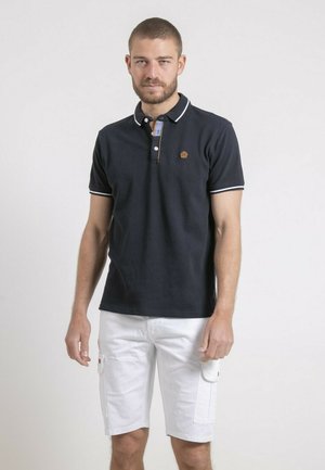 Homme portant un polo noir à manches courtes avec des bordures blanches et un short cargo blanc, debout devant un fond clair uni.