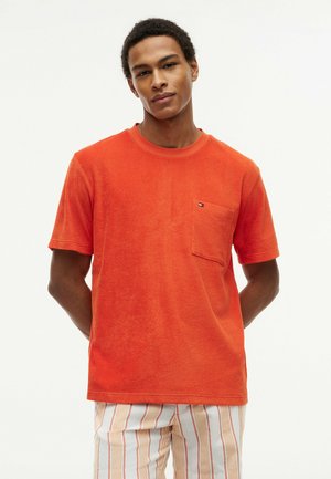 Jeune homme portant une chemise à manches courtes orange vif avec une poche poitrine et un pantalon clair rayé, debout les mains derrière le dos.