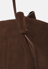 Brązowa zamszowa torba na ramię o miękkiej teksturze, złożona w unikalny sposób i z skórzanym uchwytem, z minimalistycznymi detalami w postaci przeszyć.