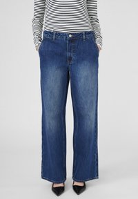 Osoba s srednjim stasom u širokim izblijedjelim plavim farmerkama, prugastoj majici s dugim rukavima i crnim cipelama s oštrim vrhom, s rukama u džepovima nasuprot jednostavne pozadine.