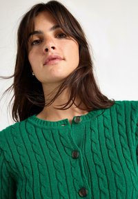 Green cable-knit cardigan with a round neckline, three dark buttons, ribbed texture, and a slight opening at the collar.