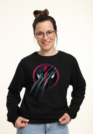 Kvinde med briller og hårrulle, der smiler, mens hun holder kanten af en sort sweatshirt med et Deadpool-logo med klo-skrabe-effekt.