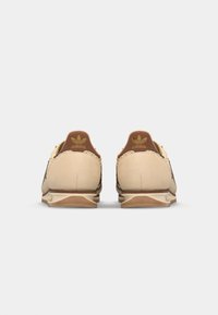 Baskets en daim beige avec un accent en cuir marron au talon, présentant une tige en maille texturée et une semelle en caoutchouc blanche. Logo doré sur le talon.