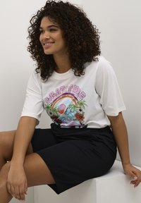 T-shirt bianco con grafica colorata "Gratitudine" che presenta palme e fiori. Pantaloni shorts neri. Tessuto liscio, vestibilità rilassata, posa seduta.