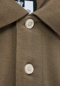 Close-up van de stof van een bruine polo, met een geribbelde kraag en twee witte knopen, waarvan er één gelabeld is met "WEEKEND," en een gestructureerd oppervlak.