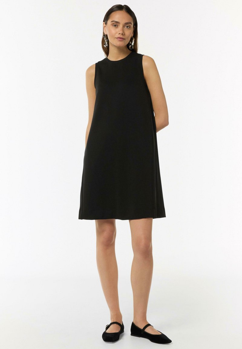 Czarna sukienka bez rękawów z okrągłym dekoltem, rozkloszowanym krojem, gładką tkaniną i długością do kolan. Nosi się z czarnymi płaskimi butami.