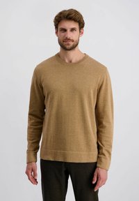 Pull en tricot beige à manches longues, col rond et poignets côtelés. Texture douce et coupe décontractée. Associé à un pantalon sombre.