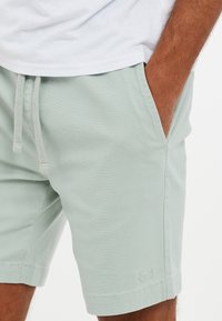 Lys grønne shorts lavet af blødt stof, med elastisk snøre i taljen, sidelommer og en diskret logo detalje på kanten.