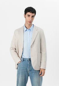Helles beige Blazer aus weichem Stoff, mit einem einzelnen Knopfverschluss und einer Tasche. Kombiniert mit einem hellblauen Button-Down-Hemd und Jeans.
