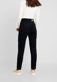 Pantalons en velours côtelé noir avec une coupe ajustée, cinq poches et une étiquette sur la ceinture. Portés avec des chaussures de couleur claire.
