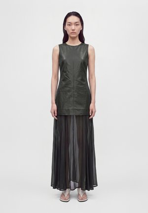 Un mannequin se tient devant un fond uni, vêtu d’un haut noir sans manches à l’aspect cuir, d’une jupe transparente longue jusqu’aux pieds et de sandales.