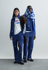 Niebieskie dresy z białymi akcentami, ozdobione gwiazdami i logo. Modelka nosi koszulkę z grafiką, a inna ma wzorzysty szal.