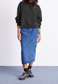 Sudadera verde con capucha y bolsillo canguro, con un logo, combinada con una falda vaquera azul. El calzado son zapatillas negras y amarillas con detalles texturizados.