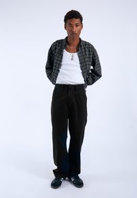 Camisa de cuadros negra sobre una camiseta blanca sin mangas, combinada con pantalones holgados negros y zapatos oscuros. Los accesorios incluyen collares en capas.
