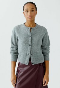 Vrouw in een grijze knooptrui en een donkerrode leren rok, staand tegen een effen witte achtergrond.
