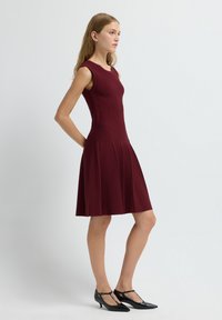 Robe sans manches bordeaux avec un corsage ajusté et une jupe évasée, présentant des plis verticaux et une texture lisse. Associée à des talons T-strap noirs.