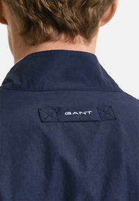 Giacca blu navy con colletto a punta, texture liscia e logo del marchio ricamato in bianco sulla parte posteriore. Dettagli cuciti sono visibili.