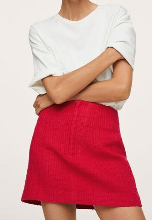 Vrouw met gekruiste armen, gekleed in een wit T-shirt met korte mouwen en een rode mini-rok met een hoog boord en textuur, tegen een neutrale achtergrond.
