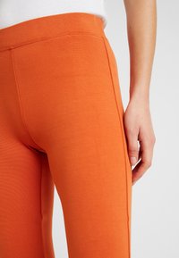 Leggings stretch orange avec une texture lisse, comportant une taille haute et un design ajusté. Détails visibles sur les coutures latérales.