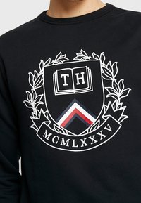 Sort sweatshirt med hvidt broderet våbenskjold med "TH", en åben bog, en laurbærkrans og "MCMLXXXV" ved bunden.