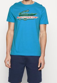 Turkosa bomull T-shirt med kort ärm, som har en grön krokodilgrafik och röd text på framsidan, ihop med mörka shorts.