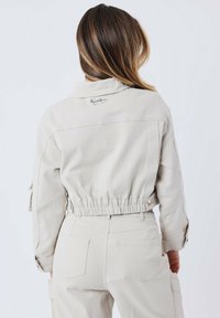 Korte beige jas met een geribbelde zoom, voorzien van een kraag en een logo detail op de rug. De stof lijkt textuur te hebben en duurzaam te zijn, met zichtbare stiksel.