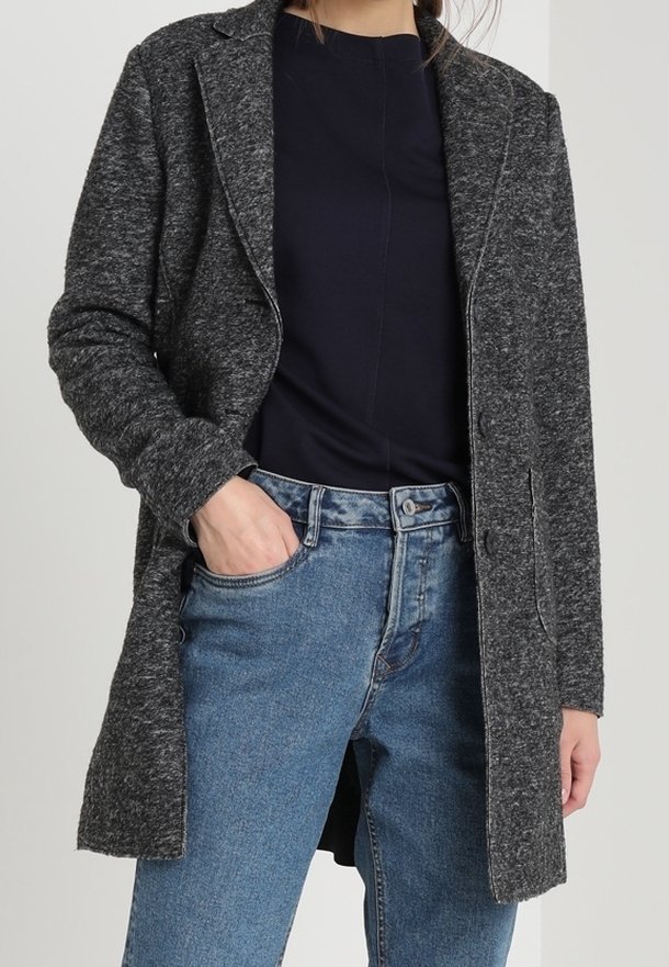 Cappotto grigio strutturato con rever a punta, due tasche frontali e taglio medio, indossato sopra una maglia scura e jeans in denim azzurro chiaro.
