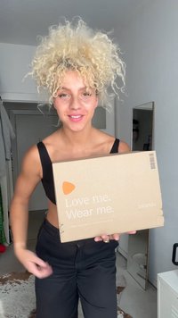 Kartono pakuotė su užrašu "Mylėk mane. Nešiok mane." balta spalva, su oranžine logotipo arka. Asmuo laiko dėžutę šviesios spalvos kambaryje.