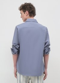 Chemise gris clair à manches longues avec manches retroussées, poignets boutonnés, col classique et texture lisse ; portée avec un pantalon rayé.