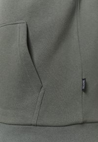 Olijfgroene hoodie van zacht materiaal, met een kangoeroe pocket, geribbelde onderkant en een klein merklabel aan de zijkant.